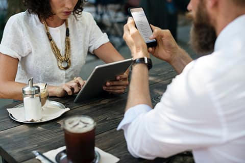 Wana-Media Mujer y hombre sentados en la mesa de un bar consultando su tablet y su teléfono móvil respectivamente